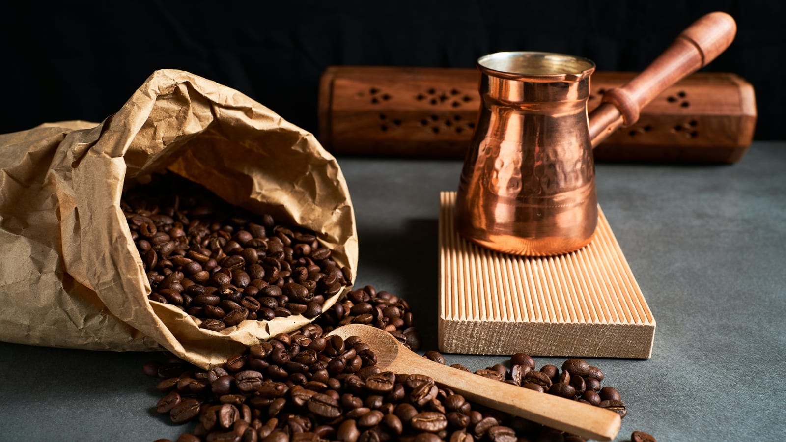 Cezve de cobre ao lado de um saco de papel com grãos de café torrados e uma colher de madeira