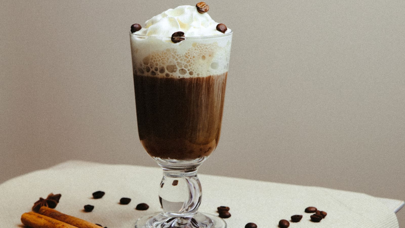Copo de irish coffee com café escuro e chantilly com grãos de café por cima, cercado por paus de canela e anis-estrelado